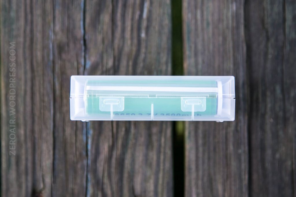 A translucent plastic case containing a green cylindrical battery is placed on a weathered wooden surface. The text ZEROAIR.WORDPRESS.COM is faintly visible along the left side.