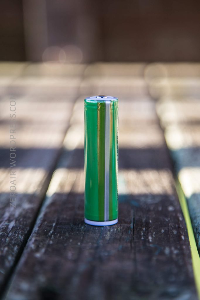 A single green and white cylindrical battery stands upright on a weathered wooden surface with a blurred background.