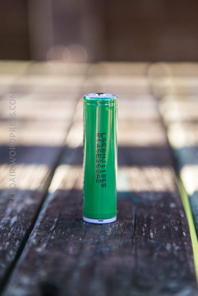 A single green cylindrical lithium-ion battery stands upright on a weathered wooden surface, with blurred background and visible text printed on the battery.