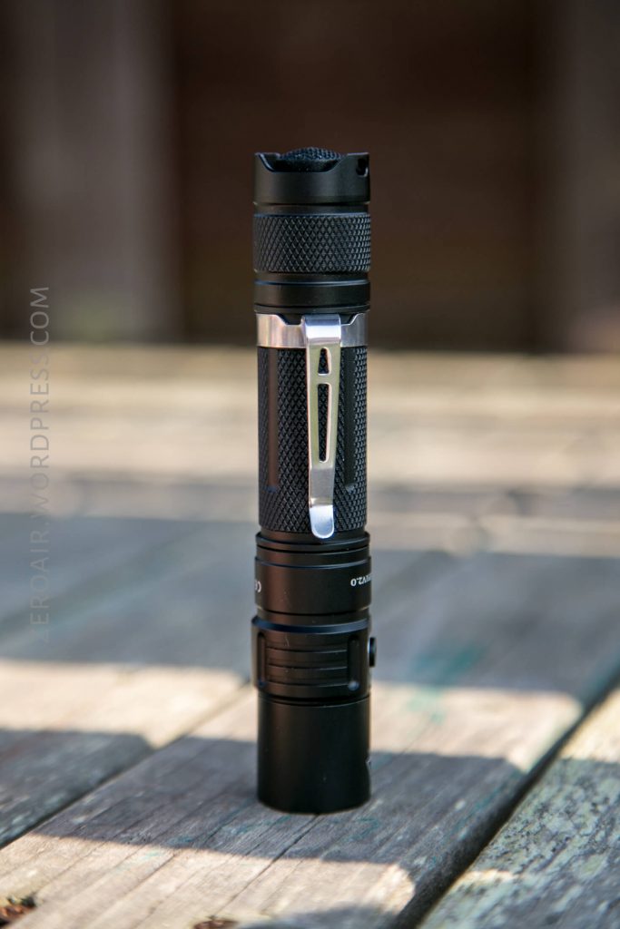 A black tactical flashlight with a metal clip is standing upright on a wooden surface. The background is blurred, and the flashlight appears to be the main focus of the image.