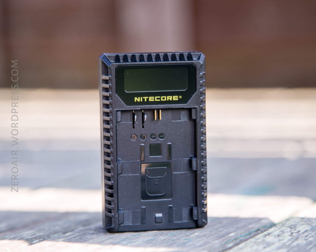 A Nitecore battery charger with a digital display and multiple charging slots is standing upright on a wooden surface. The background is blurred.