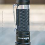 A close-up view of a black Sofirn SP12.0 flashlight, showing its textured grip, side metal clip, and part of the power button, with a blurred background.