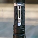 A close-up view of a black textured flashlight with a silver metal pocket clip attached to its side, shown against a softly blurred background.