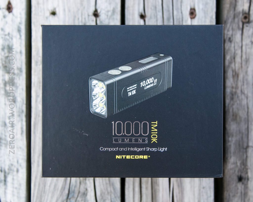 A black box displaying an image of a Nitecore TM10K flashlight with “10,000 Lumens,” product details, and branding printed on the front, is placed on a weathered wooden surface.