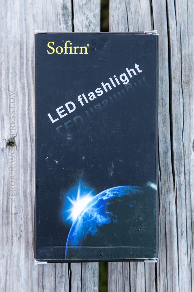 A Sofirn LED flashlight box sits on a weathered wooden surface. The box displays the brand name, product name, and an image of Earth with a bright light source near its edge.