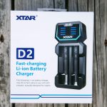 A box for the XTAR D2 fast-charging Li-ion battery charger is shown on a wooden surface. The packaging features an image of the charger, product name, and brief features in blue and black text.