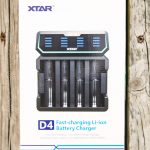A box of the XTAR D4 fast-charging Li-ion battery charger is displayed upright on a wooden surface. The box shows an image of the charger, product name, and description for lithium-ion batteries.