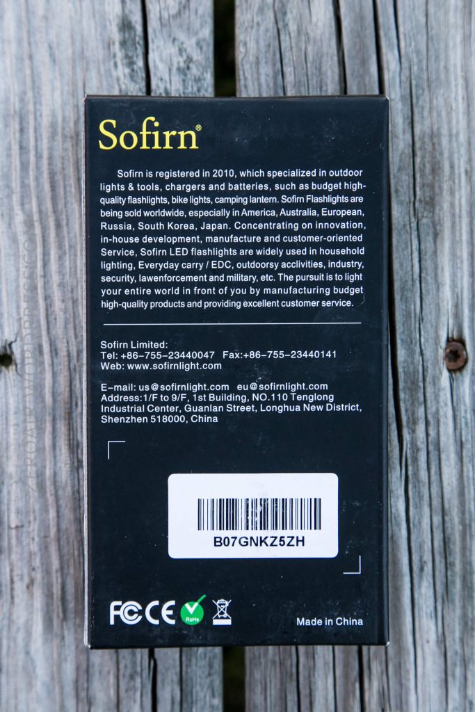 The back of a Sofirn flashlight box is shown on a wooden surface. The box features a product description, company contact information, barcode, and various certification logos.