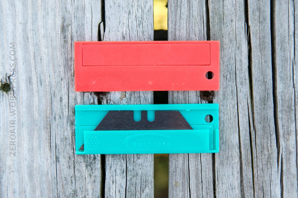 A red rectangular plastic case and a teal plastic case containing a box cutter blade are placed side by side on a weathered wooden surface.