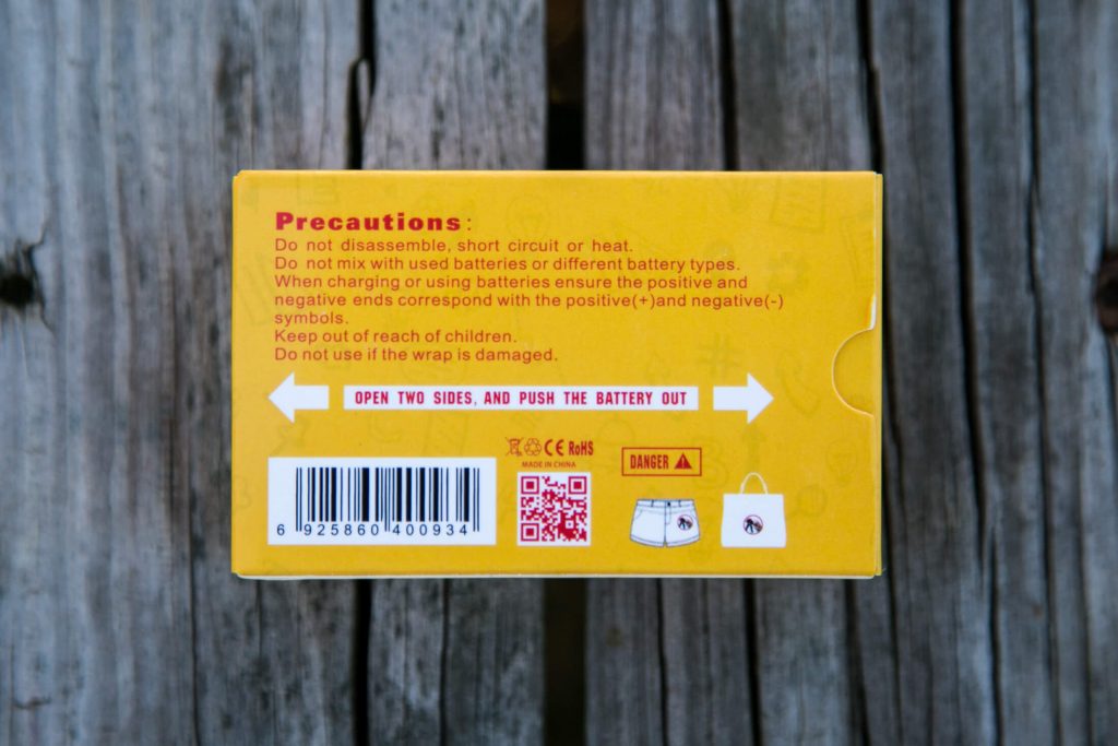 A yellow battery packaging box with red and black printed precautions about usage, including warnings against disassembly and heat, and instructions for battery installation. The box also displays barcodes and QR codes.