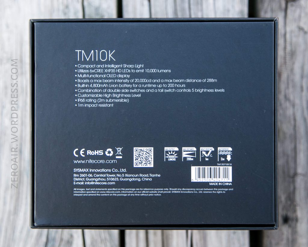 Back of a Nitecore TM10K flashlight box displaying product features, specifications, company information, certification icons, barcodes, and website URL. Text includes details about light output, beam distance, and battery life.