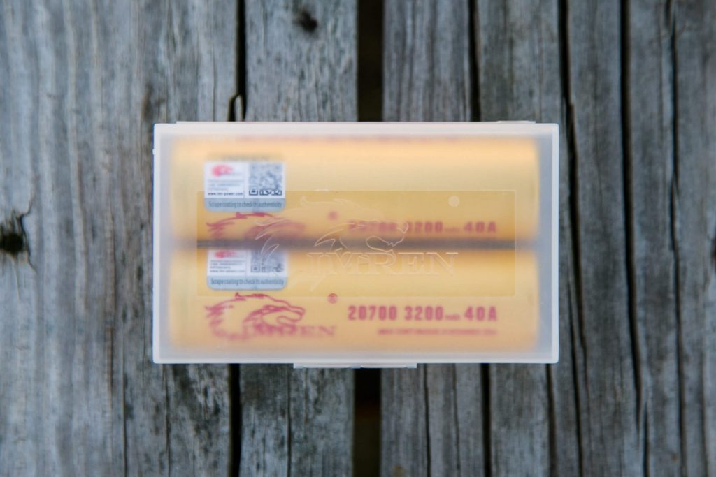 A clear plastic case holds two yellow batteries with red text and a lion logo, placed on a weathered wooden surface. The batteries are labeled 20700 3200mAh 40A.