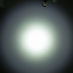 A bright white flashlight beam shines on a wall, creating a circular pattern with a soft gradient. The words ZEROAIR.WORDPRESS.COM are written vertically on the left side. Two light fixtures are partially visible at the top.