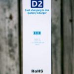 A close-up of a white box labeled D2 Fast-charging Li-ion Battery Charger, compatible with 3.6V/3.7V Li-ion/IMR/INR/ICR 18650 batteries, showing charging icons and compliance markings, on a wooden surface.