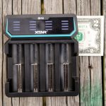 A battery charger labeled XTAR D4 holds four cylindrical batteries. A two-dollar bill is placed beside the charger on weathered wooden boards.