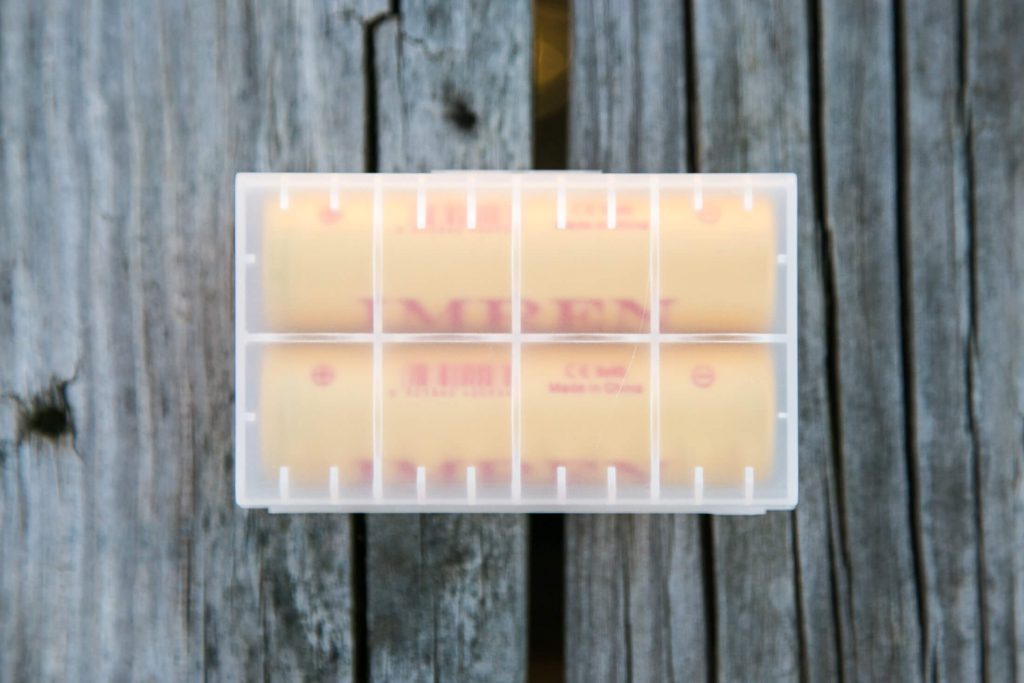 A translucent plastic case containing two cylindrical yellow batteries sits on a weathered wooden surface.