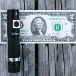 A black Sofirn flashlight is placed on top of a U.S. two-dollar bill, both resting on a weathered wooden surface.