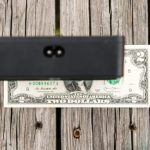 A two-dollar bill is placed on a weathered wooden surface, partially covered by a black rectangular object at the top of the image.
