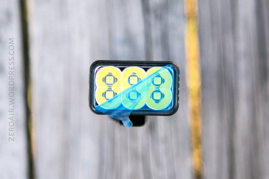 A close-up view of a rectangular flashlight head with six LED bulbs, partially covered by a piece of blue protective film. The background is wooden planks with visible grain. The words ZEROAIR.WORDPRESS.COM are along the left side.