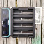 A black XTAR battery charger with six empty slots sits on a weathered wooden surface. A two-dollar bill is partially visible underneath the right side of the charger.