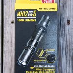 A yellow and black box for the Nitecore MH12GTS flashlight is displayed on a wooden surface. The packaging highlights 1800 lumens, USB rechargeability, and a 5-year warranty. The flashlight is shown on the front.