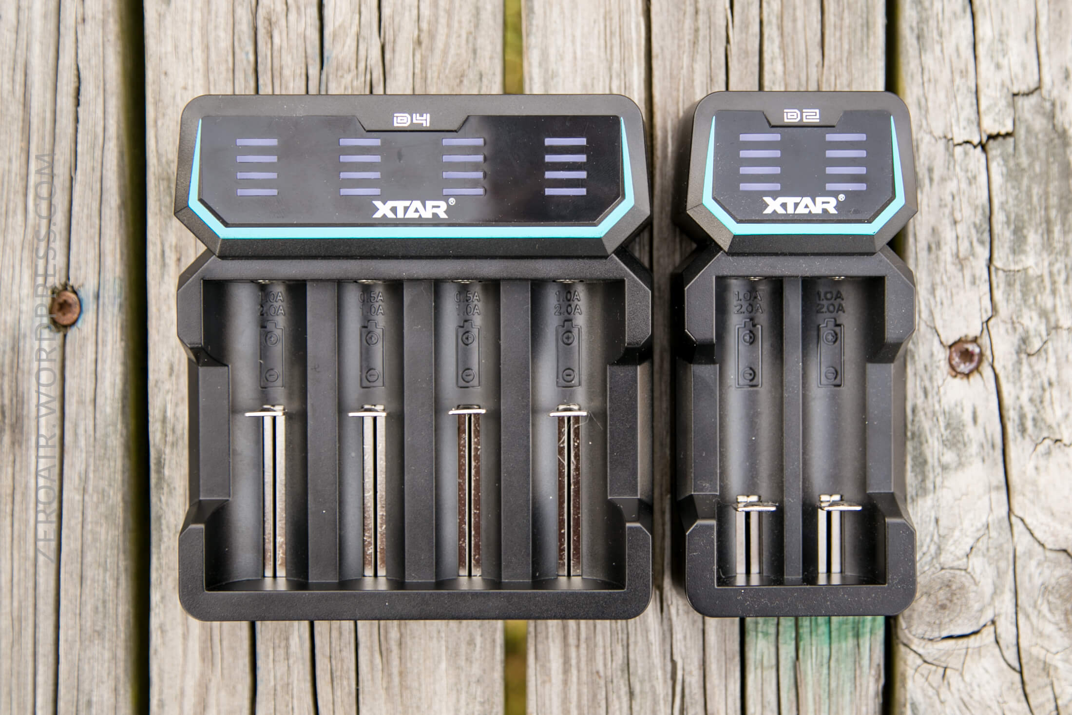 A large four-slot XTAR battery charger and a smaller two-slot XTAR battery charger are placed side by side on a weathered wooden surface. The chargers are empty and display digital screens at the top.