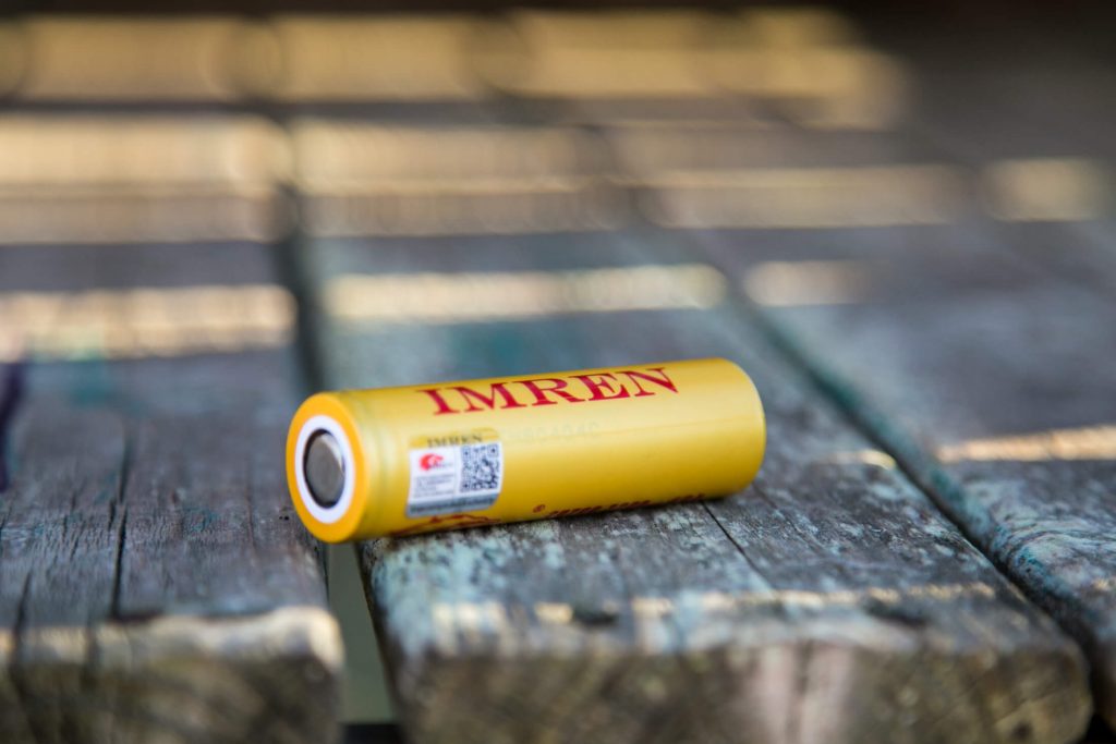 A yellow cylindrical IMREN battery lies horizontally on a weathered wooden surface, with the brand name and label visible.