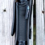 A black flashlight in a fitted fabric holster with a Velcro flap, placed vertically on a weathered wooden surface. The holster has a visible tag and a printed URL along its left side.