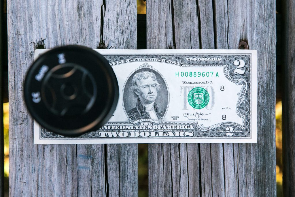 A two-dollar bill lies flat on a weathered wooden surface. A round, black camera lens partially obstructs the left side of the bill, casting a shadow.