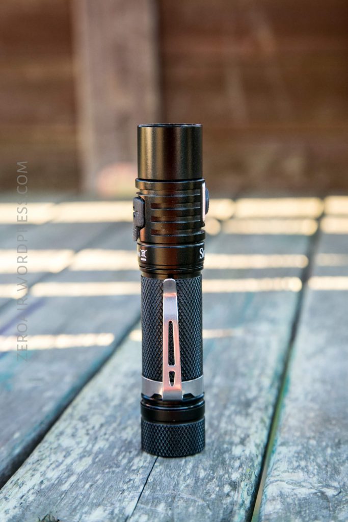 A black and silver handheld flashlight stands upright on a weathered wooden surface with sunlight casting shadows and a blurred brown background.