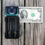 A black XTAR D2 battery charger with two batteries inside is placed on a wooden surface next to a U.S. two-dollar bill for size comparison.