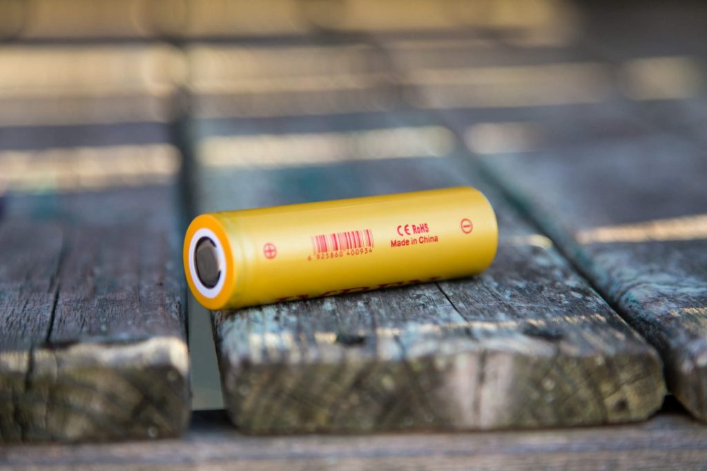 A yellow cylindrical battery with red text and a barcode is lying on a weathered wooden surface. The text includes “CE,” “RoHS,” and “Made in China.” The batterys terminals are visible on each end.