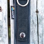 A black nylon flashlight holster with a metal snap button and a silver D-ring is attached to a weathered wooden surface. A tag with the brand name Rofis is visible on the side.