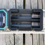 A black XTAR battery charger with two slots is placed on top of a two-dollar bill, all resting on a weathered wooden surface.