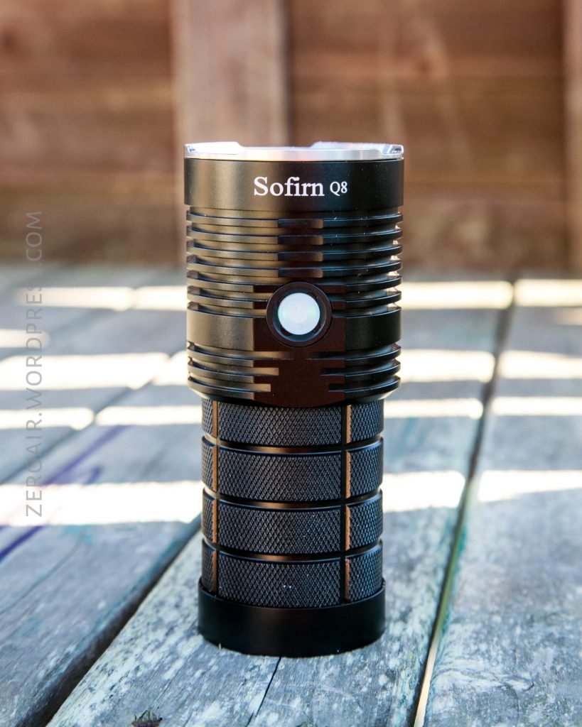 A black Sofirn Q8 flashlight stands upright on a weathered wooden surface with a blurred wooden fence in the background.