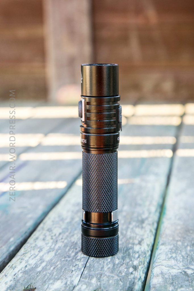A black tactical flashlight stands upright on a weathered wooden surface outdoors. The flashlight has a textured grip and visible button, with sunlight casting shadows in the background.