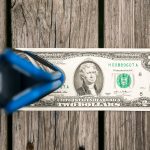 A close-up of a two-dollar bill placed on a wooden surface with a golf club head positioned above it, partially obscuring the left side of the bill.
