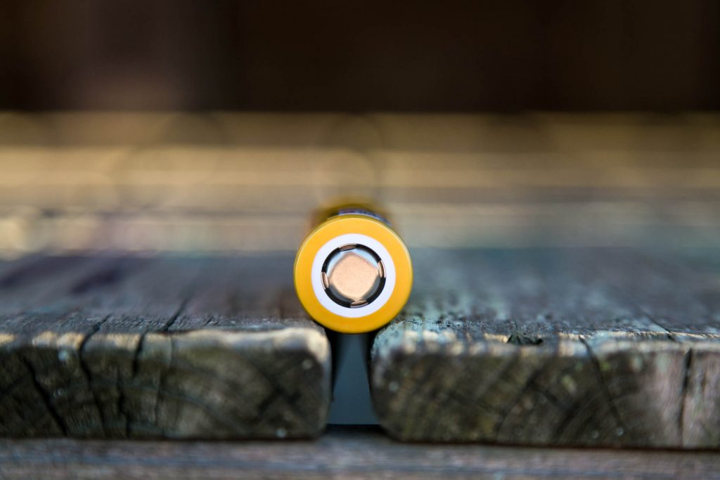 A close-up view of the flat, positive end of a yellow cylindrical battery placed horizontally between two weathered wooden planks.