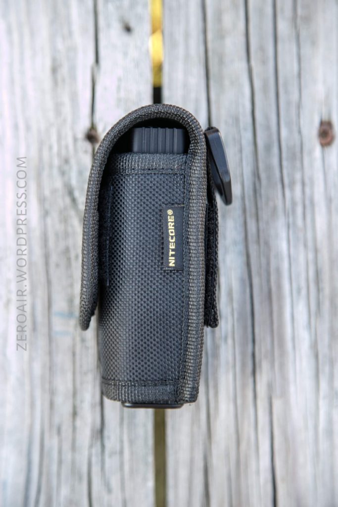 A black Nitecore flashlight in a textured fabric holster, standing vertically against a weathered wooden background. The holster has a flap closure and the NITECORE logo is visible on the side.