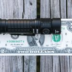 A black handheld flashlight is placed horizontally on top of a U.S. two-dollar bill, both resting on a weathered wooden surface.