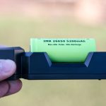 A hand holds a black battery charger with a green IMR 26650 5200mAh rechargeable battery inserted. The background is out of focus with green and brown tones.