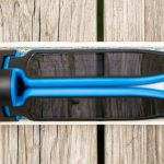 A close-up view of a blue and black sunglass lens cleaner resting on top of a rectangular object on a wooden surface. The image is taken from above.