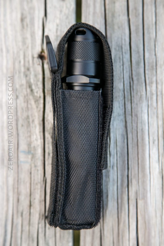 A black flashlight in a textured black nylon holster sits upright on a weathered wooden surface. The holster is open at the top, exposing the flashlight’s head.
