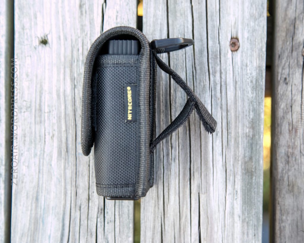 A black Nitecore flashlight in a textured holster with a Velcro flap, attached to a weathered wooden surface. The holster is upright, showing the flashlight’s end and Nitecore logo on the side.