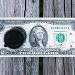 A United States two-dollar bill lies flat on a wooden surface with a round black object partially covering the left side of the bill.