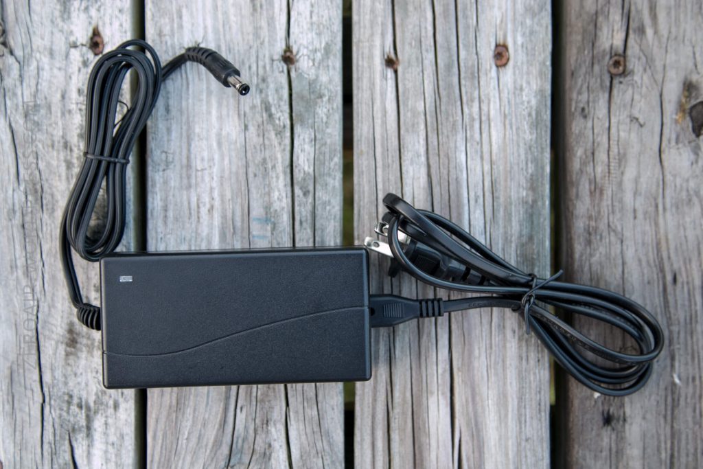 A black AC power adapter with attached cables is placed on a weathered wooden surface. The adapter has a rectangular shape with slightly rounded edges and a subtle line design on its surface.