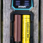A close-up of an XTAR battery charger holding two cylindrical batteries, one brown and one yellow. The yellow battery is labeled Nitecore 3400mAh 18650, and the charger is shown from above.