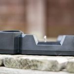 A black charging dock with a single metal charging pin is placed on a weathered wooden surface. The background is out of focus, showing brown and beige tones.