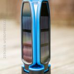 A blue and black portable solar-powered device with double-sided solar panels stands upright on a wooden surface. The background is blurred. ZEROAIR.WORDPRESS.CO is faintly visible on the left side.