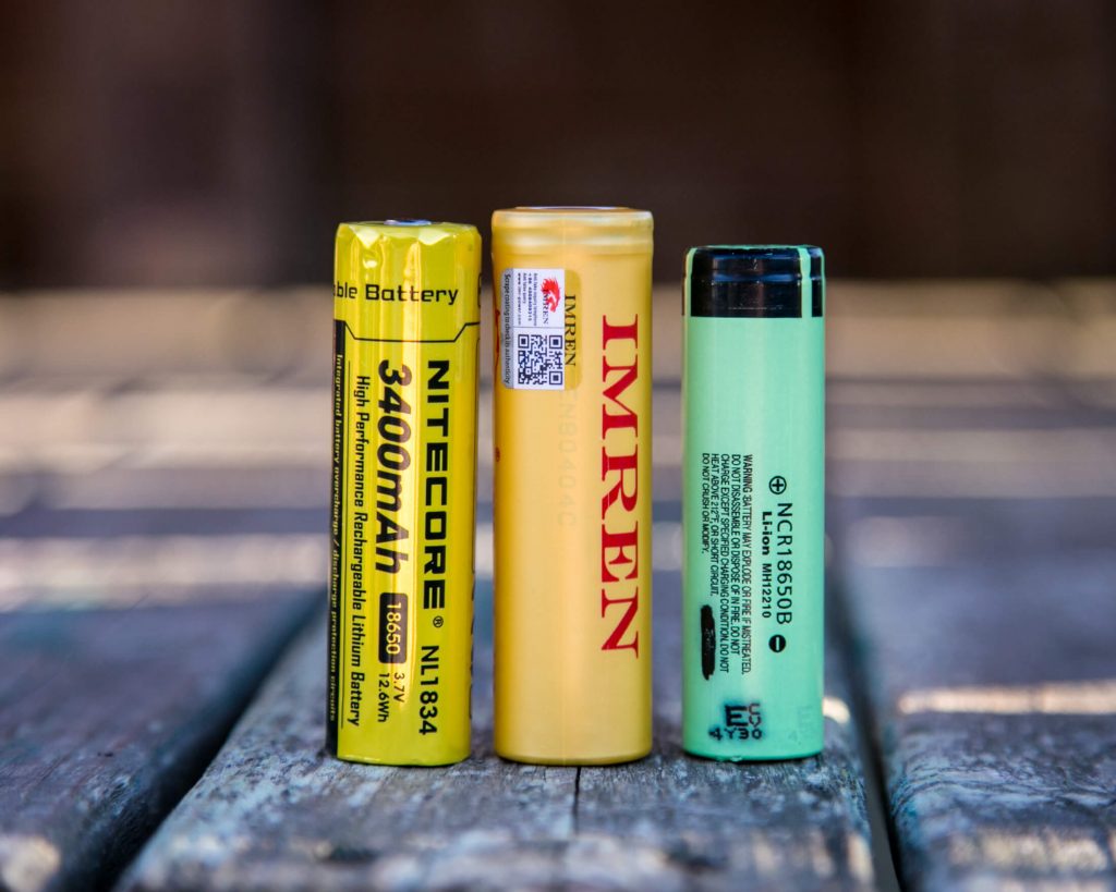Three cylindrical rechargeable batteries of different brands and colors—yellow Nitecore, yellow Imren, and green NCR—are standing upright on a weathered wooden surface.